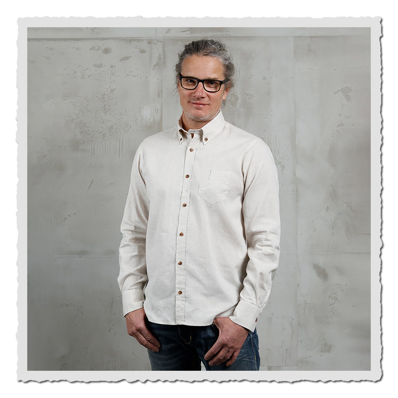 Mann steht frontal im naturweißen Hemd, Knopfleiste sichtbar, grauer Betonhintergrund.